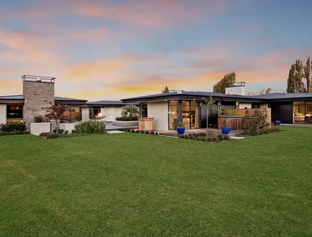 Single-storey contemporary residential home viewed from a broad landscaped lawn at dusk. The low horizontal building has dark flat roof planes, light rendered walls, stone-clad chimney volumes, extensive glazing, timber screening, and a sheltered central entry or living area. Mature trees and carefully planted garden beds surround the house.