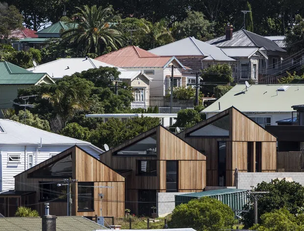A residential suburban streetscape showing a contemporary cluster of timber-clad houses in the foreground, with angular gabled forms, dark-framed glazing, and pale masonry accents. Traditional weatherboard homes with varied pitched metal roofs and mature palm and garden planting surround the newer development on the hillside.