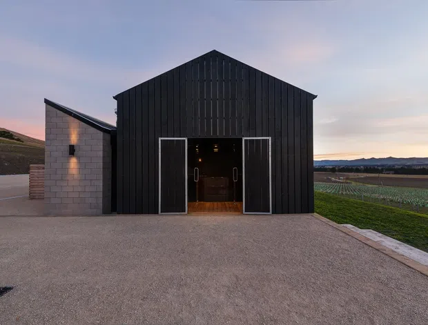 A freestanding, barn-like single-storey building is shown from the exterior at dusk. The main facade has dark vertical timber cladding, a simple gabled roof, and large central glazed double doors. A pale concrete-block side wing with a low sloping roof is attached on the left. The building sits on a broad paved forecourt beside cultivated rural fields, with distant hills visible beyond.