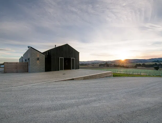 A low-rise rural building complex photographed at sunset, with a dark vertical timber-clad gabled volume beside a pale concrete-block wing. Large paved gravel forecourts surround the building, while cultivated fields, rolling hills, and distant mountains form the background. The simple shed-like massing and agricultural setting create a restrained contemporary rural character.