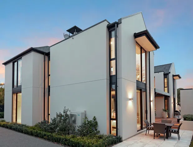 A contemporary multi-storey residential building viewed from the exterior at dusk. The design features clean white rendered wall planes, dark vertical window and door frames, extensive floor-to-ceiling glazing, projecting roof eaves, and a paved outdoor dining terrace with built-in planting around the perimeter.