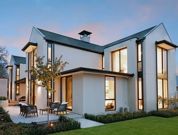 Exterior view of a large contemporary two-storey residence with crisp white rendered walls, dark pitched gabled roofs, tall black-framed windows, and projecting flat-roofed elements. The landscaped front garden includes manicured lawn, hedges, paving, and an outdoor seating terrace. Warm interior lighting glows through the extensive glazing at dusk.