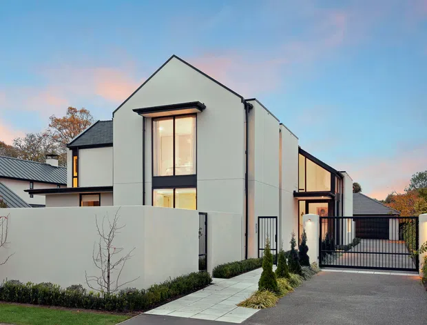 A contemporary multi-level residential home viewed from the street at dusk. The facade combines crisp white rendered walls, dark-framed windows and doors, and a steep gabled roof form. A high privacy wall, landscaped garden beds, paved entry path, and gated driveway frame the modern house.