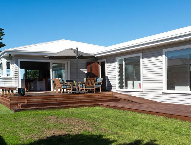 Single-storey coastal-style house viewed from the rear garden, featuring white horizontal weatherboard cladding, a low-pitched roof, large windows with white frames and shutters, and a broad dark timber deck with outdoor seating. The deck connects an open living or dining area to the lawn, while a tall evergreen tree and clear blue sky form the background.