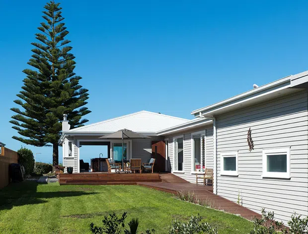Single-storey coastal-style residential house viewed from the landscaped backyard, featuring light horizontal weatherboard cladding, a pale low-pitched roof, broad windows, and a timber deck with outdoor seating beneath a covered patio. A tall Norfolk Island pine and lawn frame the exterior.