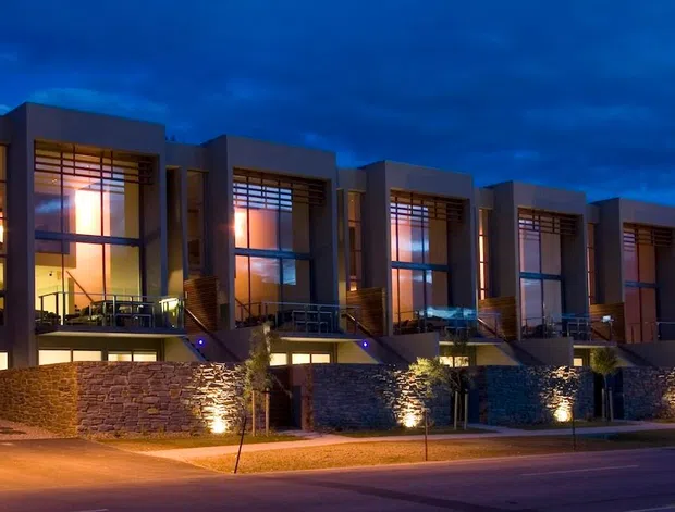 A dusk exterior view of a row of contemporary multi-storey attached residences. The buildings feature strong rectangular volumes, dark rendered facades, extensive floor-to-ceiling glazing, projecting upper-level balconies, warm interior lighting, and textured stone retaining walls with landscaped street frontage.
