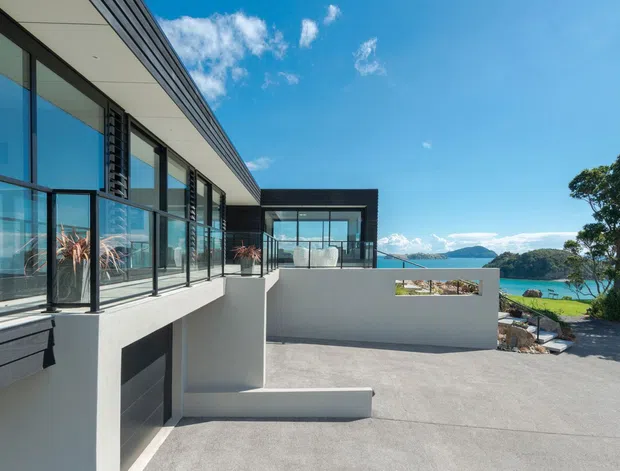 Contemporary multi-level coastal residence viewed from an elevated exterior terrace or driveway. The building features dark horizontal cladding, extensive floor-to-ceiling glazing with dark frames, crisp white rendered walls, and broad concrete paving. A turquoise bay, headland, and sky are visible beyond the property, while a small landscaped area and outdoor seating zone appear at the right.