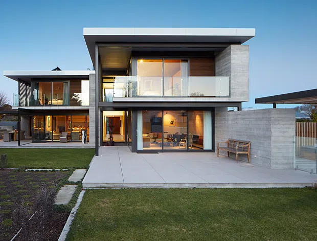 Twilight exterior view of a contemporary two-storey residence with strong horizontal lines, flat roof planes, extensive glazing, concrete or pale masonry walls, and timber-lined soffits. The house opens onto a paved terrace and lawn, with a separate open-sided carport or covered parking structure to the right. The landscaped front yard and neighbouring trees frame the architectural composition.