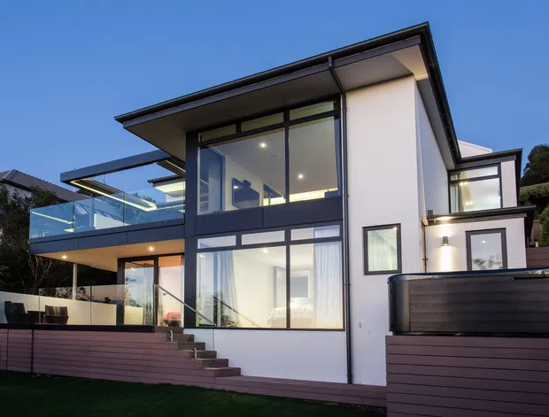 Exterior view of a contemporary multi-level residence at dusk, featuring crisp white rendered walls, dark metal-framed floor-to-ceiling glazing, broad roof overhangs, a cantilevered upper terrace with clear glass balustrades, timber-clad lower walls, and an elevated spa or pool enclosure at the right.