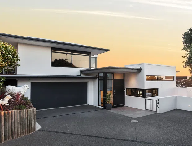 Street-facing view of a contemporary two-storey residence with a flat roof, white rendered walls, dark aluminium-framed glazing, a large dark garage door, and a recessed entry. The home has a broad paved driveway, low white boundary walls, and a timber-plank raised planter with established planting.