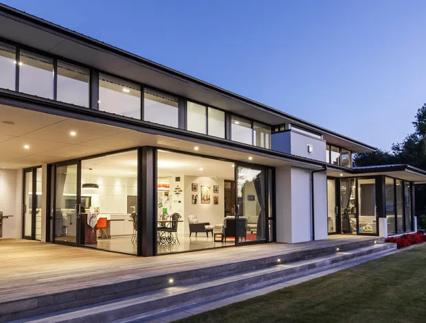 Twilight exterior view of a contemporary two-storey residence with a long low-pitched roofline, extensive floor-to-ceiling glazing, dark window frames, white rendered walls, and a broad timber terrace accessed by wide steps. Warm interior lighting reveals open-plan living and dining areas through the glass, with landscaped lawn and garden edges in the foreground.