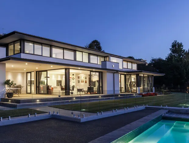 Twilight exterior view of a large two-storey contemporary home with broad floor-to-ceiling glazing, projecting flat roof planes, white rendered walls, dark framing, and a landscaped lawn beside a swimming pool. Warm interior lighting is visible through the glass, with a glass pool safety fence in the foreground.