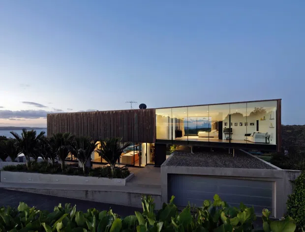 Exterior twilight view of a modern multi-level residence with a flat roof, extensive floor-to-ceiling glazing, a timber-clad upper volume, concrete retaining walls, and an integrated garage. Tropical planting surrounds the foreground, while a distant coastal horizon is visible beyond the property.