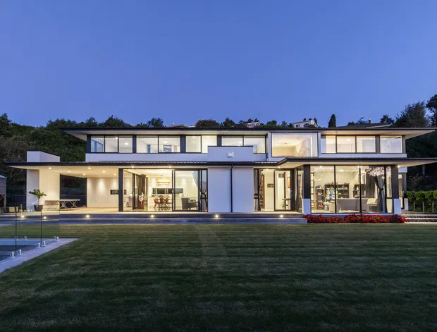 A wide dusk exterior photograph of a large two-storey contemporary residence with a low, nearly flat roofline, extensive floor-to-ceiling glazing, white rendered walls, and dark horizontal framing. The house opens onto a broad manicured lawn and a long narrow swimming pool, with illuminated interiors and subtle garden planting visible around the facade.