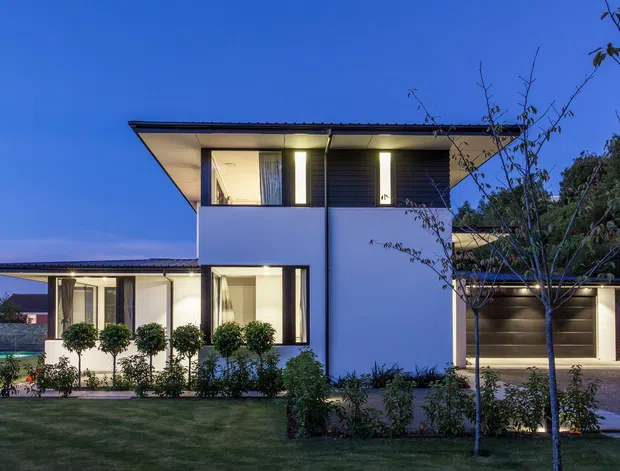 Twilight exterior view of a contemporary two-storey residence with crisp white rendered walls, dark horizontal cladding, broad roof overhangs, large glazed windows, and an integrated garage. The house is set within a landscaped suburban garden, with warm interior lighting visible through the windows.