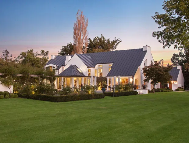 A large white residential house viewed from an expansive manicured lawn at dusk. The home features multiple gabled roof forms with dark standing-seam metal roofing, white rendered walls, projecting bay-like glazed areas, a covered entrance or veranda, and warm interior lighting. Mature trees and hedges surround the property.