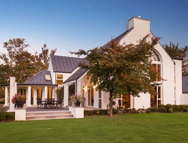 Exterior view of a large white residential home with steeply pitched standing-seam metal roofs, prominent chimney-like vertical forms, tall arched glazing, and a covered outdoor terrace. The landscaped grounds include a manicured lawn, clipped hedges, mature trees, and planted entry columns. The home has a refined traditional architectural character with contemporary detailing.