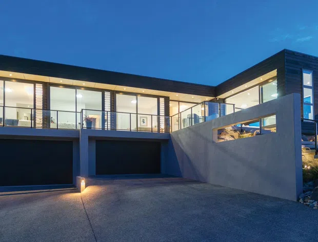 Contemporary multi-level residential house viewed from the driveway at dusk. The home features dark horizontal cladding, extensive floor-to-ceiling glazing, strong geometric forms, integrated double garage doors, a concrete retaining wall, and illuminated interiors and exterior steps. The site appears elevated and sloped, with exposed rock landscaping.