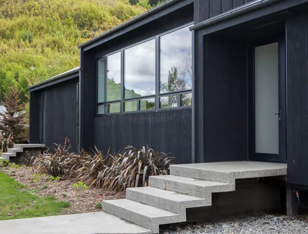 Exterior view of a contemporary dark-clad building set against a steep green hillside. The structure features vertically ribbed black cladding, large aluminium-framed windows, a recessed entry door, and a projecting concrete stairway leading from a gravel and planted garden approach. A smaller connected or adjacent volume is visible to the left.