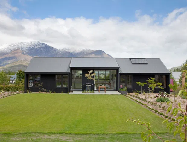 Single-storey contemporary dark-clad house with a long, low gabled roof and extensive glazed central living area, set in a carefully landscaped garden with a broad lawn. Snow-capped mountain ranges form the background.