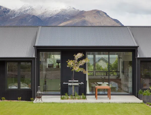 Contemporary single-storey dark-clad house with a broad corrugated pitched roof and extensive floor-to-ceiling glazing. The central glazed section opens onto a concrete terrace with a small table and planter, while mountain peaks and a grassy lawn form the setting. Dark vertical cladding, charcoal-framed windows and doors, and a simple linear facade create a restrained alpine architectural character.