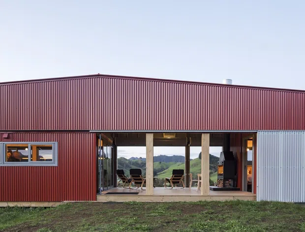 A low, single-storey building with a broad gently sloping roof and corrugated metal cladding. The main facade is deep red corrugated metal, with a contrasting pale grey corrugated section at the right. Large open glazed doors reveal a sheltered living area with timber posts, chairs, and a fireplace or dark built-in element, while a compact horizontal window punctuates the left wall. Rolling green hills are visible through the opening.
