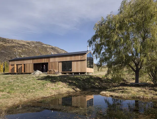 Single-storey contemporary timber-clad house set in a rural mountainous landscape beside a reflective pond. The long, low building has a simple pitched roof, dark-framed glazing, timber doors, and a prominent glazed gable end. Natural grasses, mature trees, and rocky hills surround the residence.