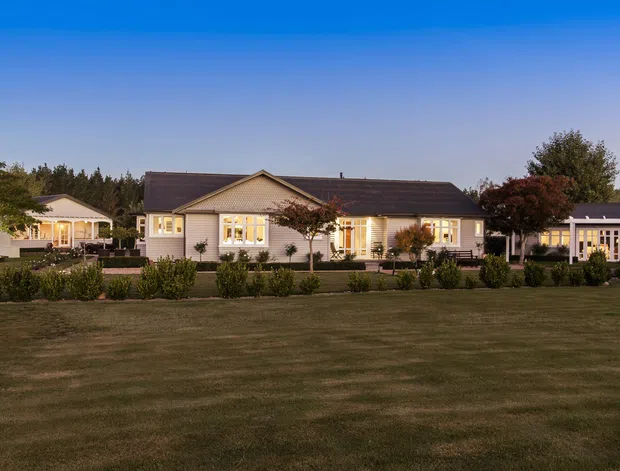A broad single-storey suburban or rural residence photographed at dusk, with a long low gabled roof, pale horizontal cladding, illuminated windows, covered verandah-like outdoor areas, landscaped garden beds, and a large open lawn in the foreground.