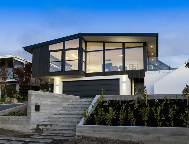 Contemporary two-storey residential facade at dusk, featuring strong geometric volumes, extensive floor-to-ceiling glazing, dark vertical cladding, a flat roof, an integrated garage, and a landscaped concrete stairway leading to the entry.