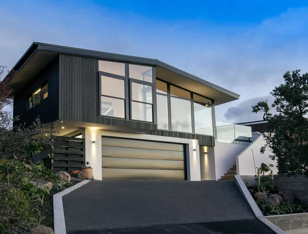 Contemporary two-storey residence viewed from the street at dusk, with dark vertical cladding, extensive floor-to-ceiling glazing, a broad low-pitched roof, and an integrated double garage. A steep asphalt driveway leads to the garage, with landscaped planting and exterior stairs along the sides.