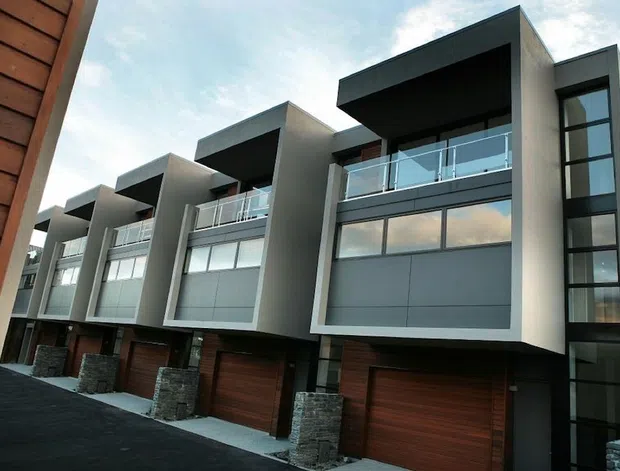 Exterior view of a row of attached contemporary multi-storey residences. The buildings feature strong geometric forms, dark grey and white rendered facade panels, large glazed openings with glass balustrades, vertical window strips, timber garage doors and timber wall cladding, plus stacked-stone elements at the entries. A shared driveway runs along the front.