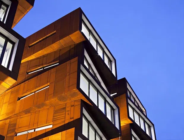 Upward-looking architectural exterior view of a modern multi-storey building with strongly projecting box-like forms, dark-framed floor-to-ceiling windows, and warm orange-brown timber or wood-toned cladding illuminated against a clear blue sky. The composition emphasises the angular facade, deep overhangs, and layered balconies or projecting volumes.