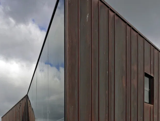 Exterior architectural photograph of a sharply angular, gable-ended building elevated on slender supports. The facade is clad in dark weathered brown metal panels with pronounced vertical seams, while the triangular end wall is largely glazed and reflective. A second matching wing recedes behind it, with a rocky landscaped ground plane, concrete retaining edge, and glimpses of a coastal-looking horizon beneath dramatic overcast skies.