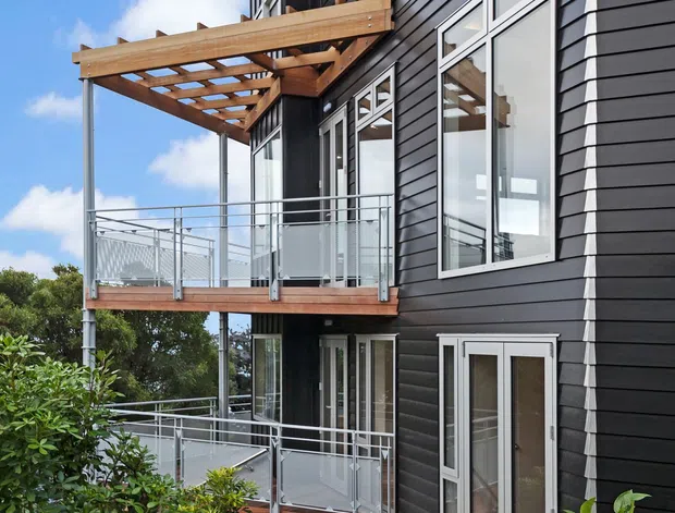 Exterior view of a multi-level house with dark horizontal cladding, white-framed windows and doors, stacked timber decks with clear metal railings, and a timber pergola. A landscaped garden and stepped timber path lead toward the building.