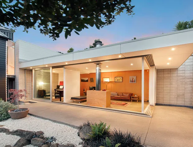 A modern single-storey flat-roof extension with extensive floor-to-ceiling glazing, opening onto a paved courtyard and minimalist landscaped garden. The interior visible through the glass includes a warm-toned living area with timber flooring, built-in seating and a compact kitchen or bar element. The exterior uses pale masonry walls, white soffits and dark cladding on the adjoining structure.