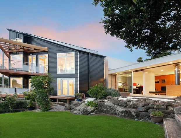 Exterior view of a contemporary multi-level residence at dusk, featuring dark horizontal cladding, a sloped metal roof, extensive glazing, a timber pergola and balcony, and a flat-roofed open living extension. The landscaped foreground includes a manicured lawn, rock garden, planting, and steps leading toward the house.