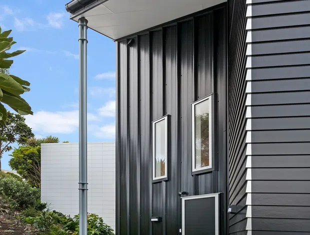 Exterior architectural photograph of a contemporary dark-clad house entrance. The facade combines vertical black metal cladding with horizontal dark weatherboard-style siding, a projecting eave with a round gutter and downpipe, narrow vertical windows, and a dark entry door. A timber deck and concrete path lead to the entrance, with landscaped planting beside the building under a clear blue sky.