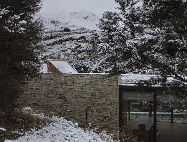 Exterior view of a low-profile building set among snow-covered trees, with a rugged natural stone wall, dark-framed floor-to-ceiling glazing, and a partially visible timber-clad pitched roof. Snowy alpine mountains rise in the background, creating a secluded mountain setting.