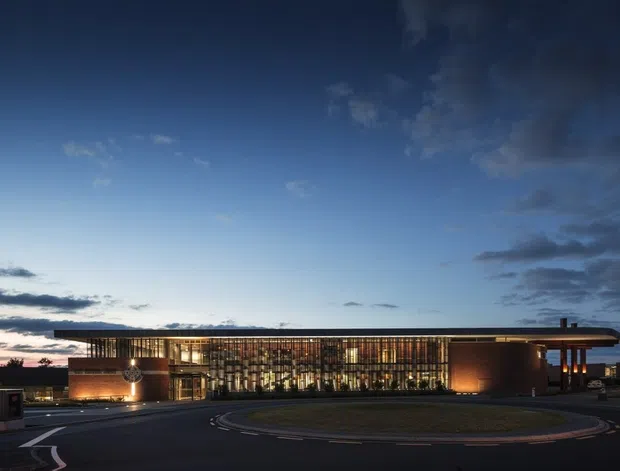 A low, contemporary commercial building photographed at dusk, with a long glazed central facade framed by warm-toned solid wall volumes and a broad projecting flat roof. The building fronts a circular drive and landscaped forecourt, with interior lighting creating a warm glow against the blue evening sky.