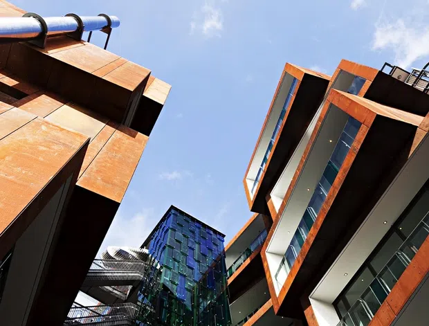 Upward-looking architectural view between several contemporary multi-storey buildings. The facades feature strong angular forms, extensive glazing, dark metal surfaces, and prominent rust-orange weathered metal cladding, with exposed rooftop and balcony structures against a bright blue sky.