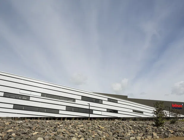 Exterior view of a long, low modern commercial building with layered horizontal white and dark facade bands, narrow ribbon windows, and a gently sloping roofline. The building sits behind a broad rocky foreground, with a visible Hellmann logo on the right side and an overcast sky above.
