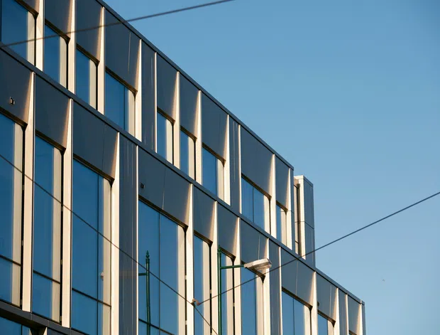 Close-up exterior view of a contemporary multi-storey building facade with a regular grid of tall reflective glazed panels, dark metal framing, and projecting vertical fins. Overhead utility wires cross the clear blue sky.