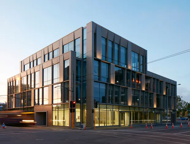 Exterior view of a contemporary multi-storey commercial building at a street intersection. The building features a rectilinear massing, extensive floor-to-ceiling glazed curtain walls, dark metal framing, and warm-toned vertical facade panels. Street infrastructure, traffic lights, power lines, and construction cones are visible in the foreground.