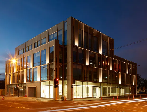 A contemporary multi-storey commercial building photographed at dusk from a street corner. The facade combines extensive floor-to-ceiling glazing with warm-toned metal or panel cladding, projecting rectangular volumes, and illuminated ground-floor frontage. Traffic light infrastructure, street markings, construction cones, and light trails frame the urban streetscape.