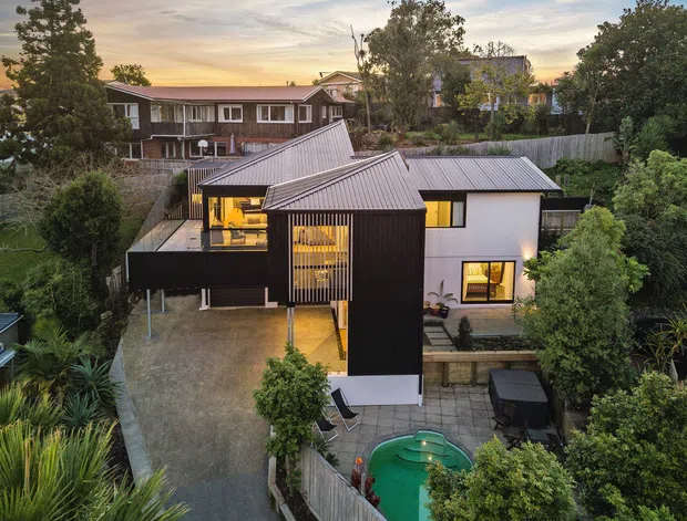 Aerial view of a contemporary multi-level residential house in a leafy suburban setting. The home combines dark vertical cladding with white rendered walls, sharply pitched metal roofs, extensive glazing, timber screening, a paved outdoor entertaining area and a small swimming pool. Established trees and neighbouring homes surround the property, with warm interior lighting visible at dusk.