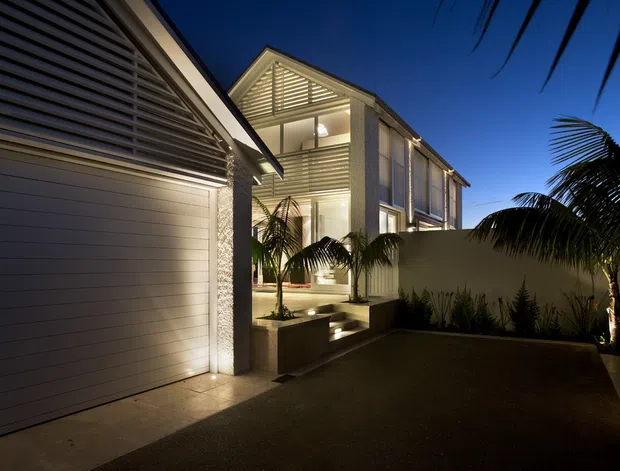 Night-time exterior view of a contemporary multi-storey residence. The composition shows a large enclosed garage with horizontal paneling at left, a recessed illuminated entry courtyard with steps and tropical planting, and a light-coloured upper-level facade featuring extensive glazing and horizontal louvres. Palm foliage frames the scene, while concealed landscape lighting highlights the walls and entry path.