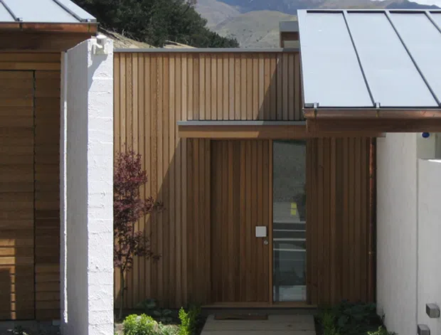 A compact modern residential entrance courtyard with a straight concrete pedestrian path leading to a timber-clad entry door. Vertical natural timber boards form the central facade, contrasted with white textured masonry walls and low-maintenance gravel landscaping. Lean-to metal standing-seam roof planes project over the entry and adjacent structures. Mountainous terrain is visible beyond the property, with established trees framing the scene.