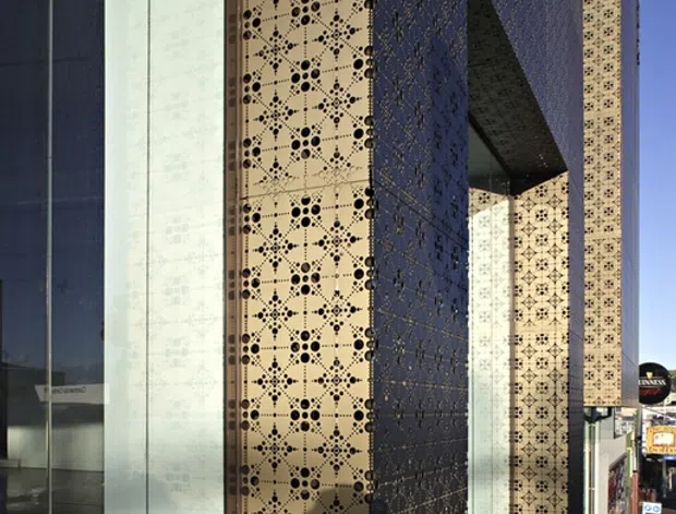 Street-level view of a contemporary commercial building facade with tall glazed panels and prominent patterned perforated metal screens. A deep projecting dark screen forms a dramatic recessed entry or opening, contrasting with cream-coloured geometric perforated panels and reflective glass.