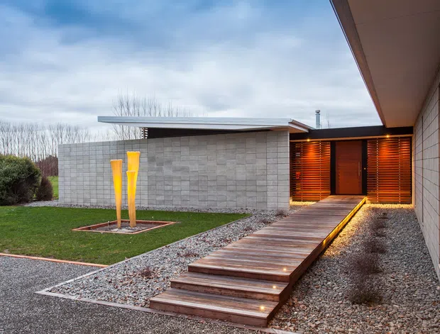 Exterior view of a low-slung contemporary building approached by a raised timber boardwalk and steps over a gravel landscape. The facade combines pale masonry block walls, warm timber louvres and a recessed timber entry, with concealed linear lighting along the walkway and sculptural outdoor lights in the lawn.
