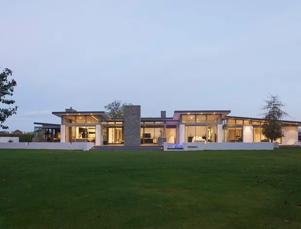 Wide exterior view of a low, single-storey contemporary residence set behind an expansive lawn. The home has broad floor-to-ceiling glazing, flat or very shallow-pitched roof planes with pronounced overhangs, warm interior lighting, white rendered walls, and a prominent central grey stone-clad feature volume.